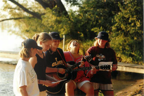 Five teenagers in the early 2000s happily sing and play guitar by the lake